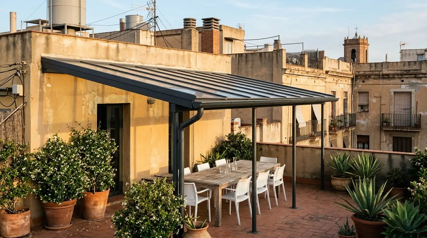 Terraza de un ático cubierta con un techo de aluminio y cristal, con mesa y sillas debajo y los tejados de Barcelona al fondo
